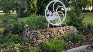 Un jardín con una escultura metálica decorativa, diversas plantas y un muro de piedra.