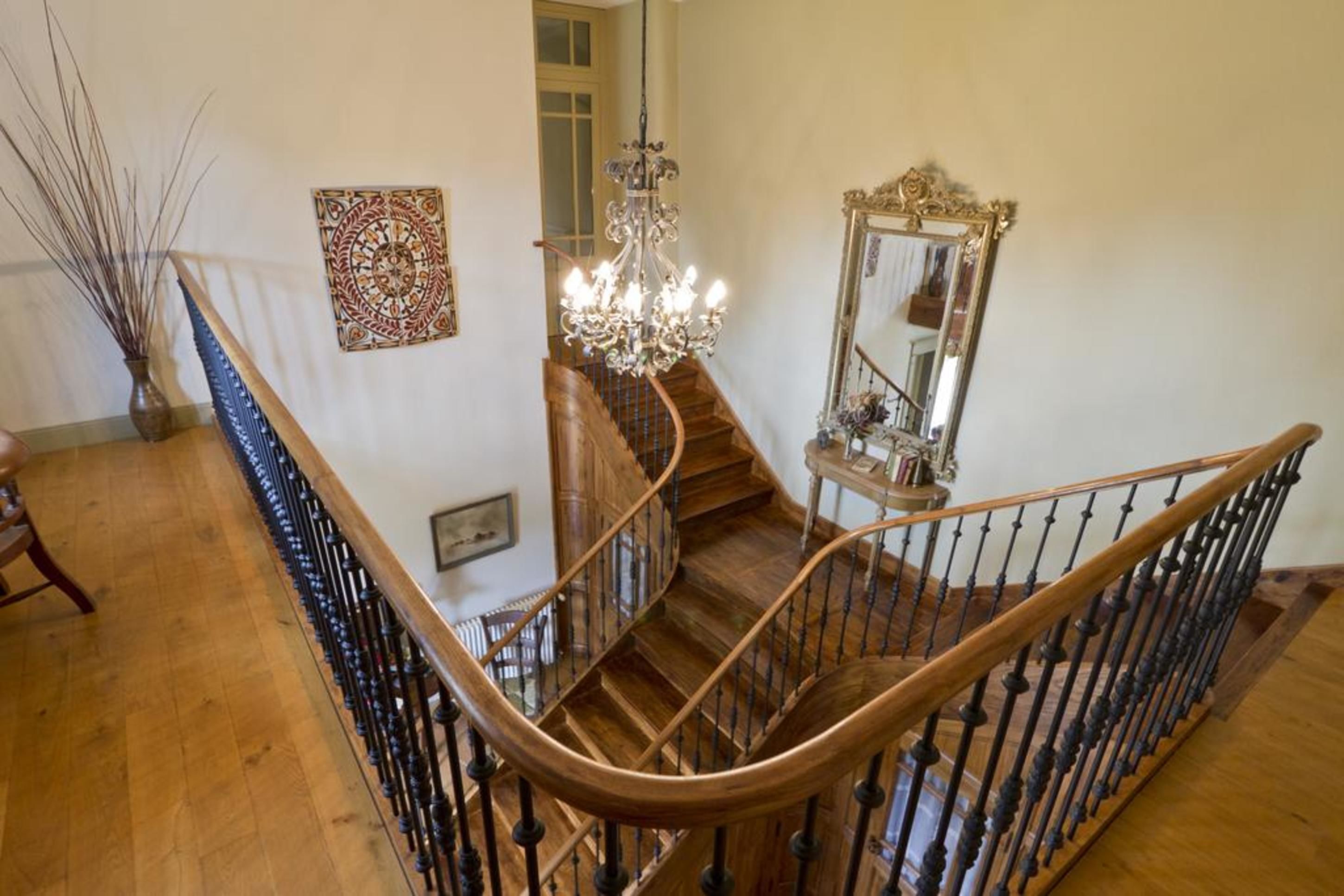 A grand staircase with a wooden handrail and black balusters, leading to a landing with a chandelier and a mirror.