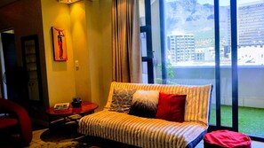 A modern living room with a sofa, a small table, and a chair. There is a large window with a view of buildings and a mountain. - Cape Town Apartment with balcony (Cape Town)