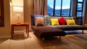 A modern living room with a sofa, a small table, and a view of a cityscape through large windows. - Cape Town Apartment with balcony (Cape Town)