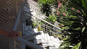 A balcony with white sun loungers, a lattice railing, and a view of a garden with pink flowers and green foliage. - Porto Assini (Nafplio)