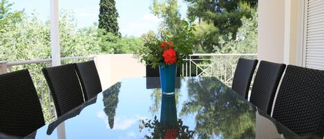 Una mesa con cubierta de vidrio, sillas de mimbre negro, un jarrón azul con flores rojas y vistas a árboles y una valla blanca.