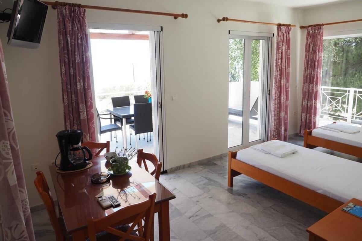 A room with a bed, a table with a coffee maker, and a chair. There is a balcony with a table and chairs outside.