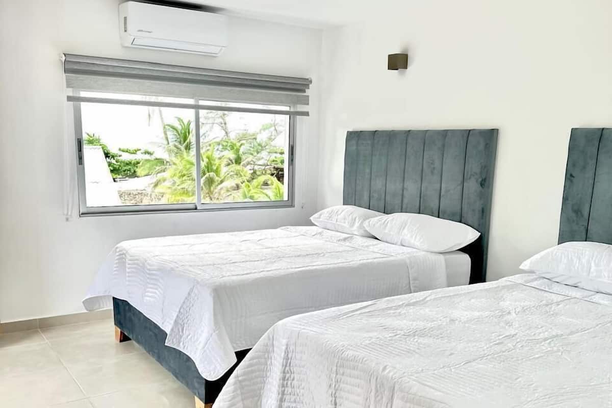 A modern bedroom with two beds, a large window with a view of greenery, and a ceiling air conditioner.