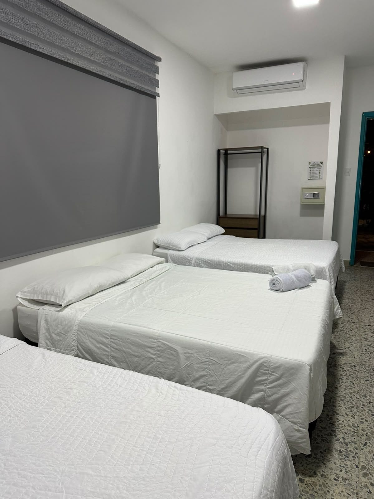 Habitación de hotel con dos camas, una ventana con persianas, un equipo de aire acondicionado, un espejo y una puerta.