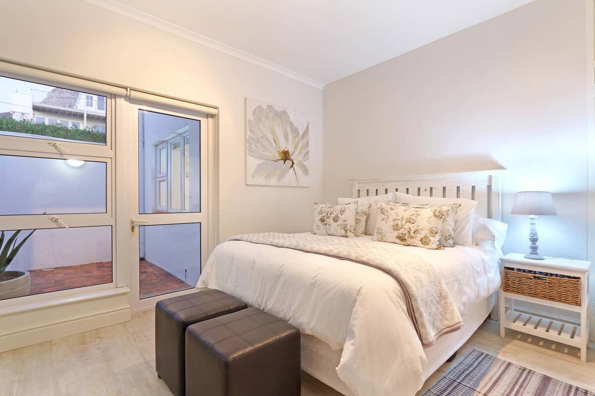 Ein Schlafzimmer mit großem Fenster, einem Bett mit floralem Bettzeug, einem Nachttisch mit Lampe und einer Topfpflanze.