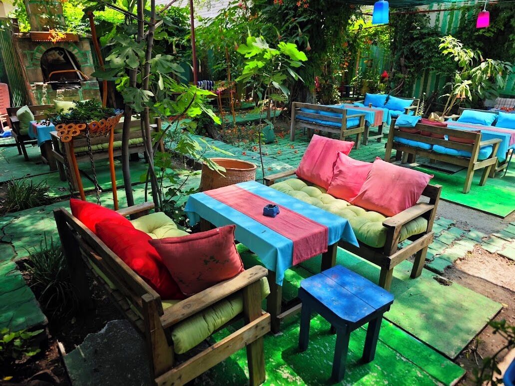 An outdoor seating area with wooden benches and tables, surrounded by greenery.