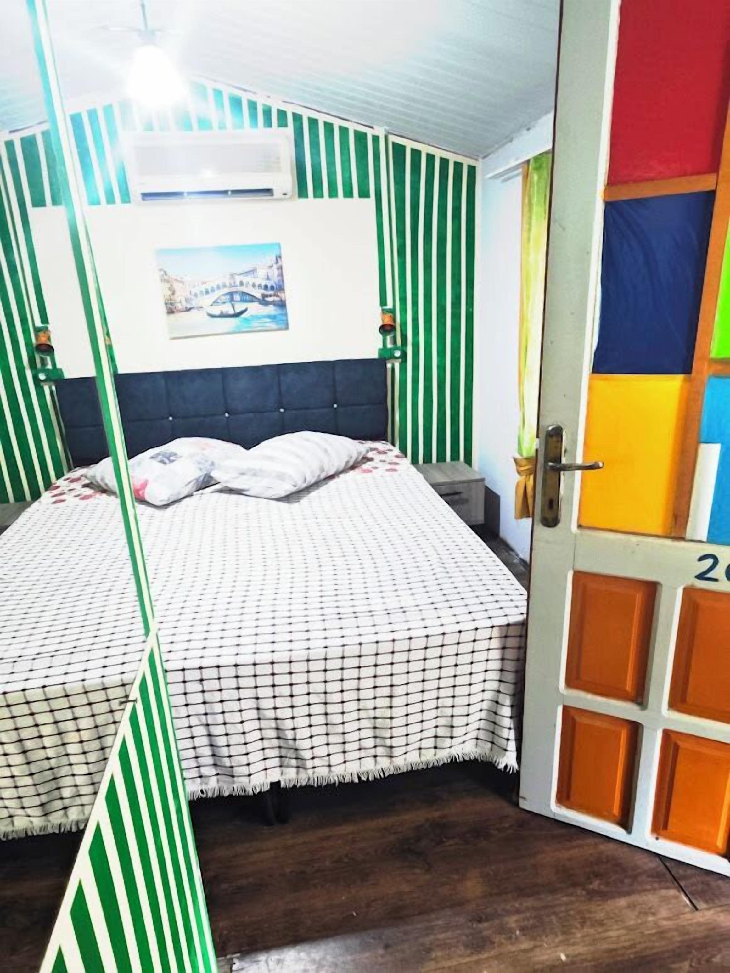 A room with a bed, a door with colorful panels, and a window with a view of a building.