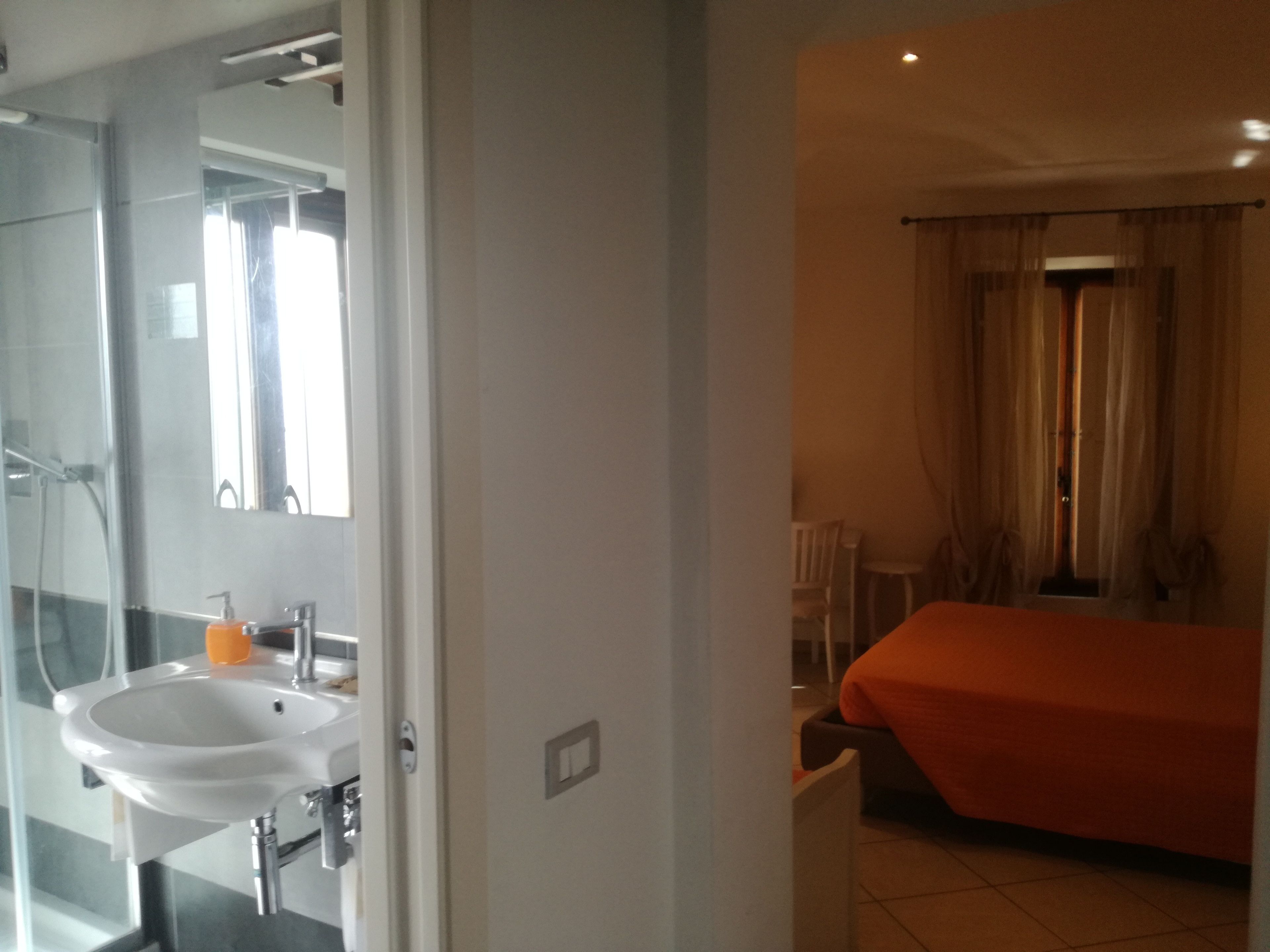 Une chambre d’hôtel avec une salle de bain comprenant un lavabo et un miroir, un lit recouvert d’une courtepointe orange et une fenêtre avec des rideaux.