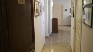 A hallway with a wooden door, white walls, and tiled flooring. There are framed pictures on the walls and a radiator on the wall. - Camollia Suites (Siena)