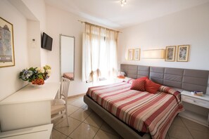 A hotel room with a bed, bedside table, chair, desk, and a TV. - Camollia Suites (Siena)