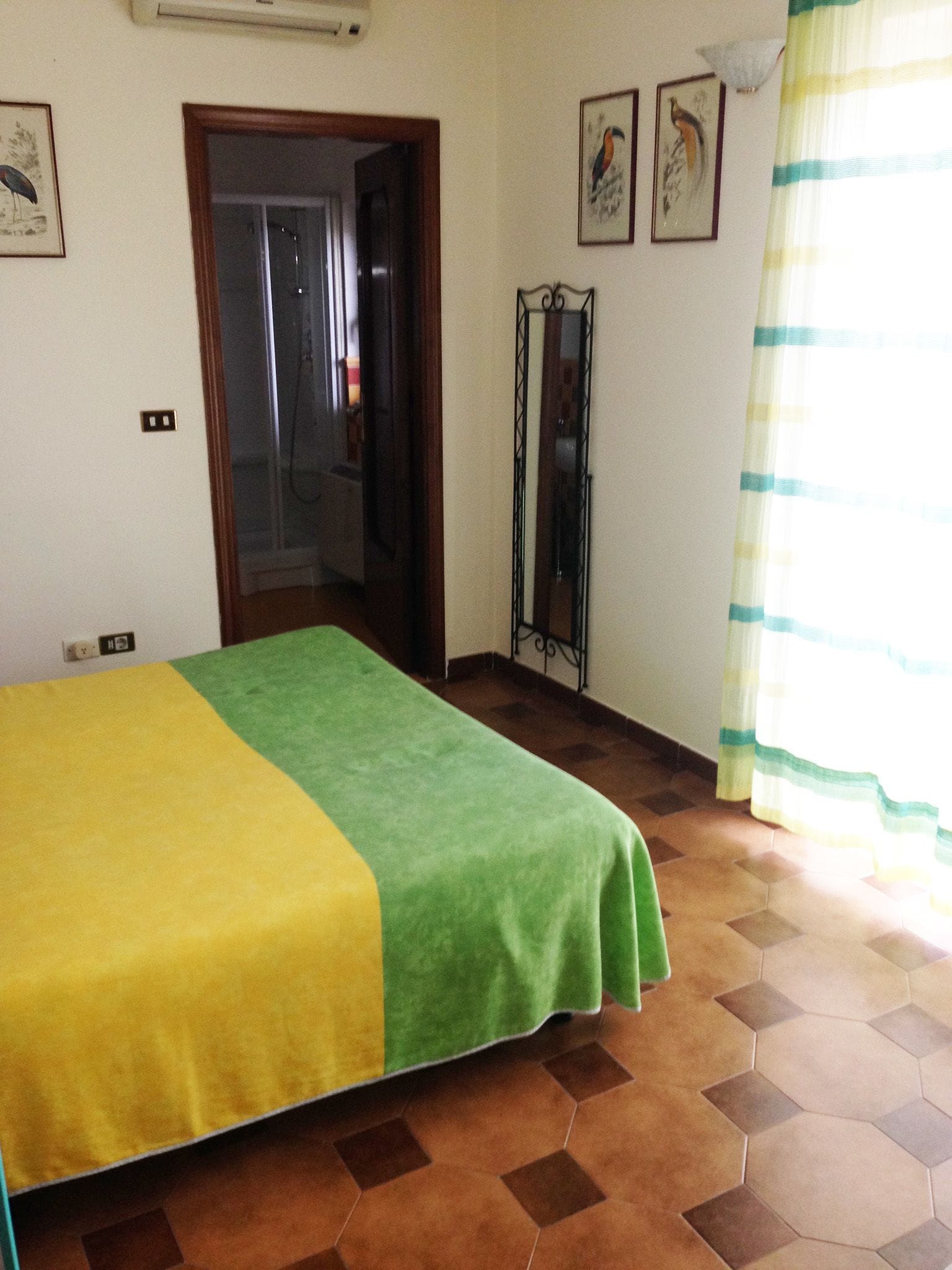 A room with a bed covered in a green and yellow blanket, a standing mirror, and a window with a striped curtain.