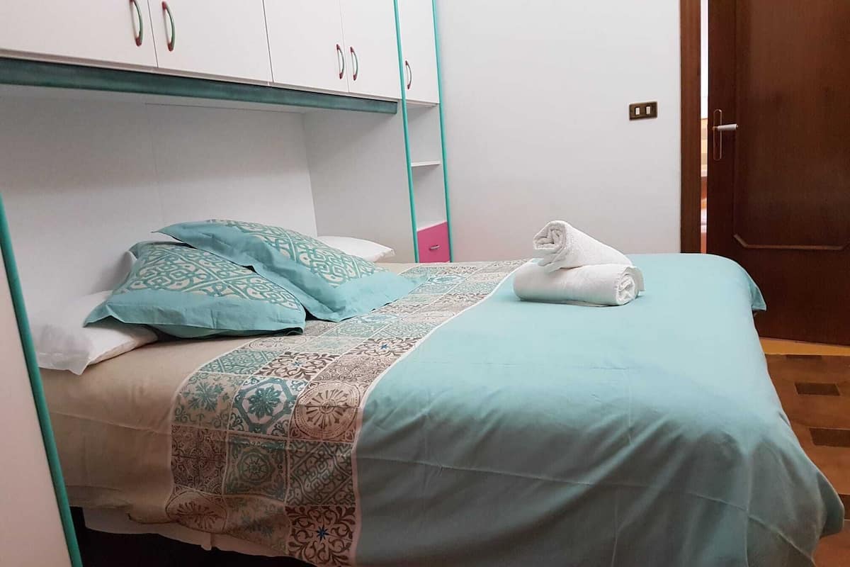 A single bed with a patterned blanket and pillows, a folded towel on the bed, and a wardrobe with white doors and teal handles.
