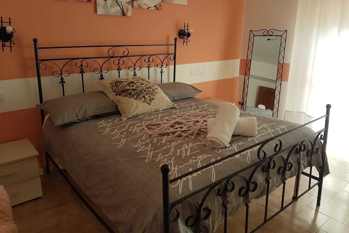 A bedroom with a metal bed frame, a single bed with a patterned blanket, a nightstand, a wall-mounted light, and a mirror.