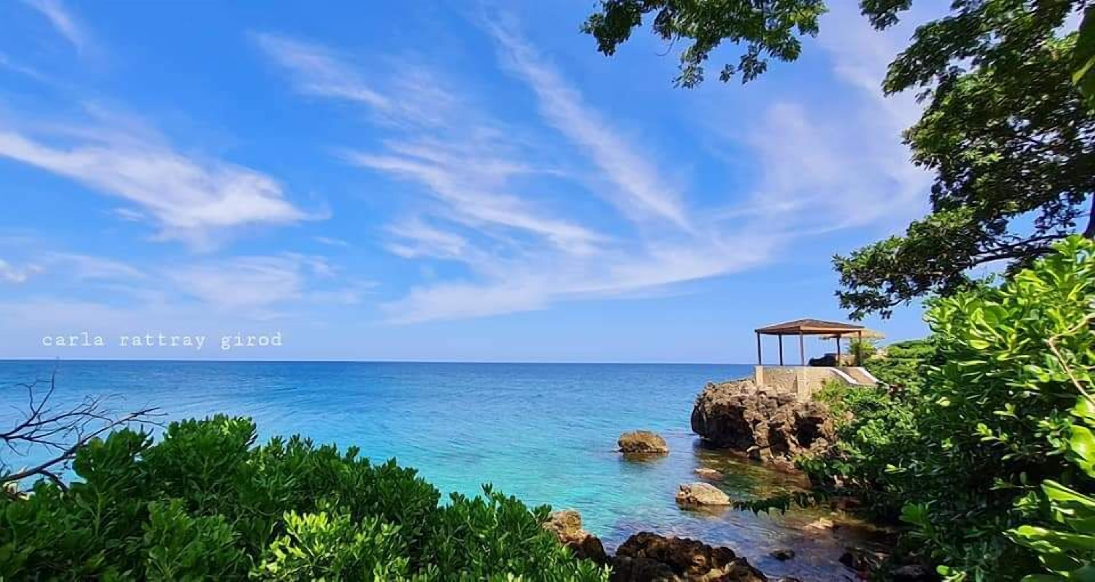 Uma paisagem costeira com um coreto sobre rochas, céu azul límpido e mar calmo.