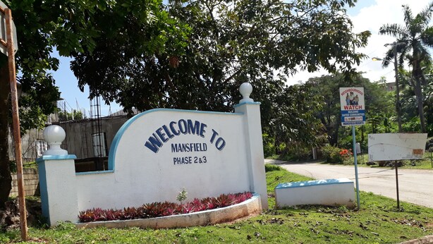 Señal de bienvenida para Mansfield Fase 2 y 3, con un área ajardinada y una señal de tráfico al fondo.
