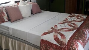 Una cama bien tendida con colcha floral, una mesita de noche con lámpara y una ventana con cortinas de gasa.