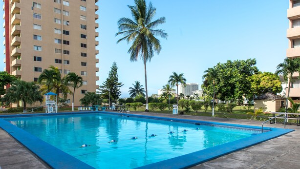 A swimming pool with clear blue water, surrounded by palm trees and a tall building in the background. - Deluxe Turtle Towers Apartments (Ocho Rios)