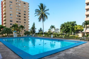 A swimming pool with clear blue water, surrounded by palm trees and a tall building in the background.