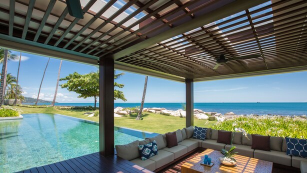 Ein moderner Außenbereich mit Holzterrasse, einer eingelassenen Couch mit gemusterten Kissen und Blick auf das Meer und den Strand.