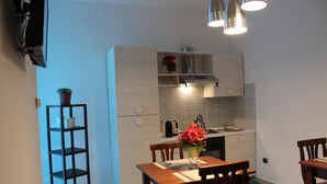 A compact kitchen with a dining area, featuring a wooden table, chairs, and a shelf with a potted plant. - Harmony Rooms (Catania)