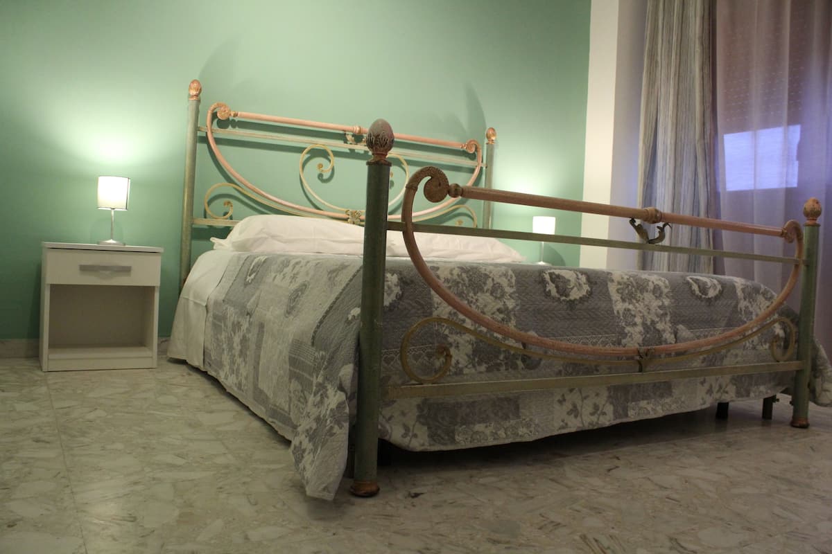 A bed with a metal frame and floral bedding, a bedside table with a lamp, and a plain wall with a curtain.