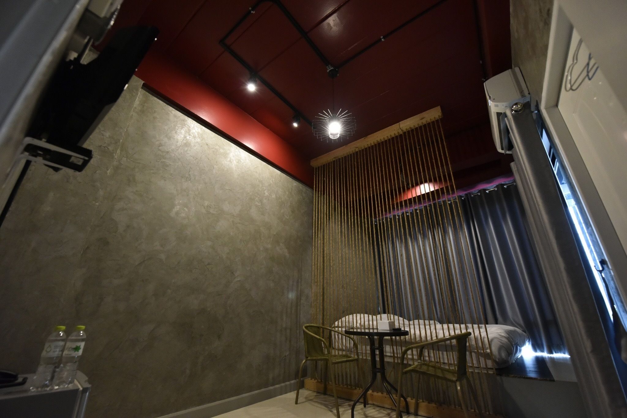 A small, dimly lit room with a bed, a chair, and a table. There is a curtain divider and a ceiling light.