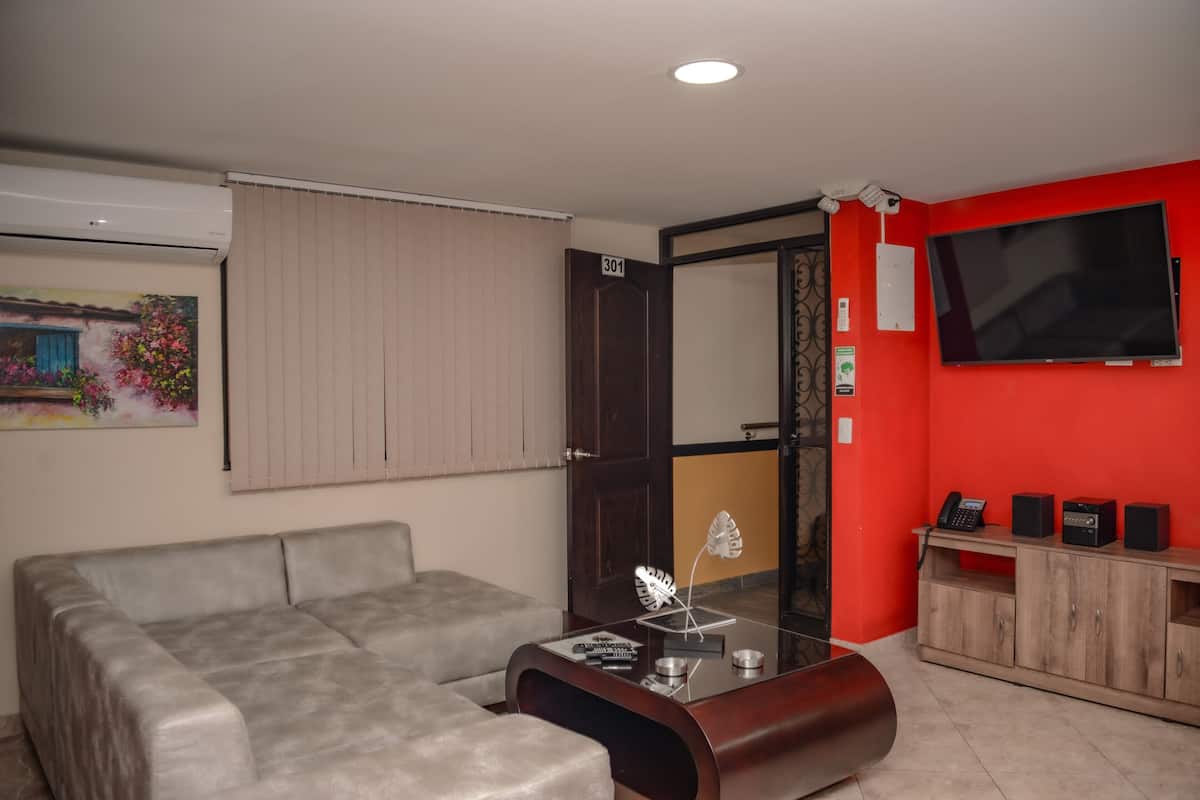 A living room with a grey sofa, a glass coffee table with decorative items, a wooden cabinet with speakers, and a red wall with a TV.