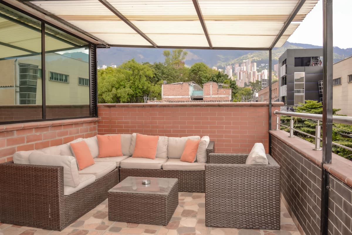 A covered patio area with a wicker sofa and a small table, overlooking a cityscape with buildings and trees.