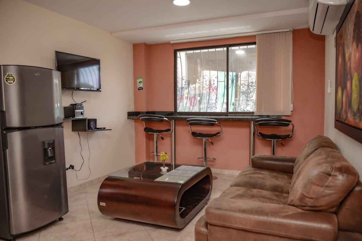 A compact living space with a kitchenette, a sofa, a coffee table, and a TV mounted on the wall.