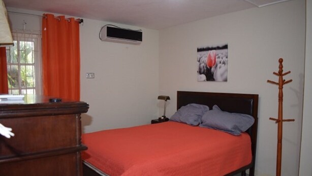 A bedroom with a bed, a bedside table, a lamp, a coat rack, and a window with curtains. - Waterloo Suite (Kingston)