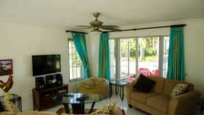 A living room with a television, speakers, a glass coffee table, a sofa, a bean bag, and curtains. - Seas the Day Sanctuary Little Bay (Negril)