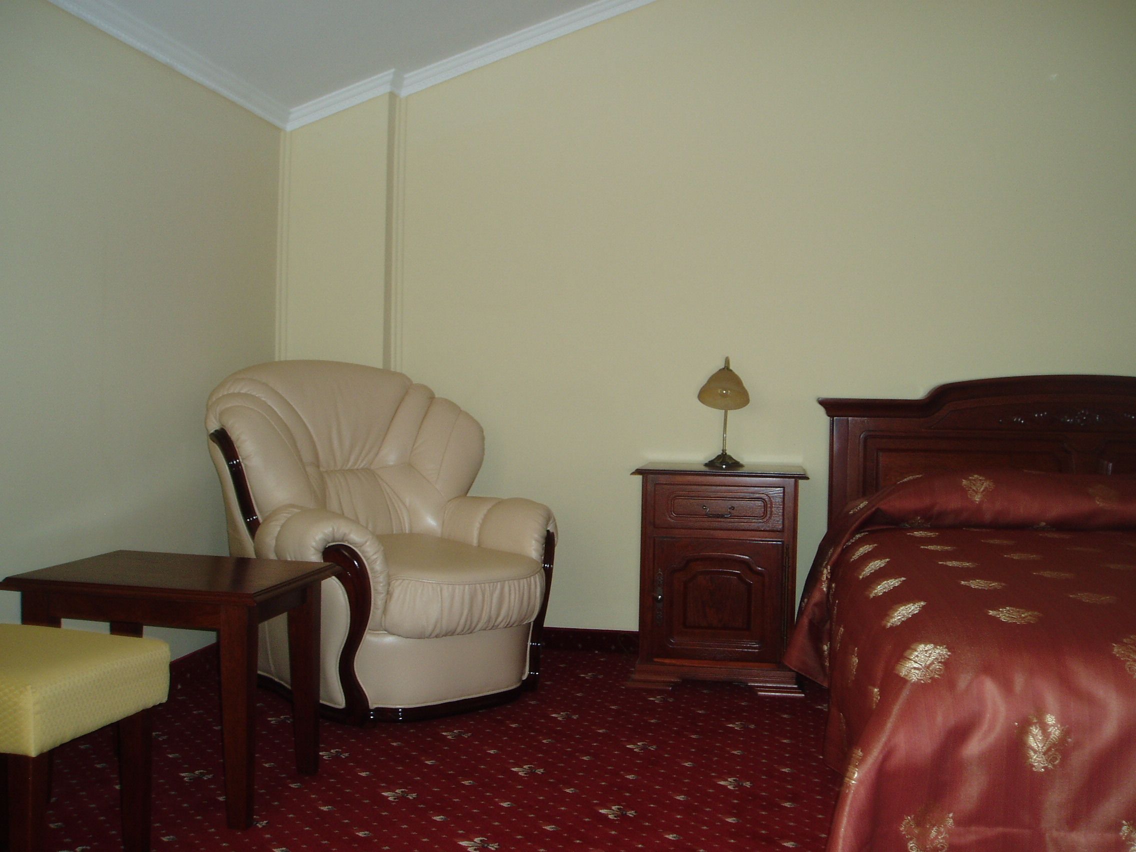 A hotel room with a large armchair, a wooden nightstand with a lamp, a bed with a red bedspread, and a small table with a chair.