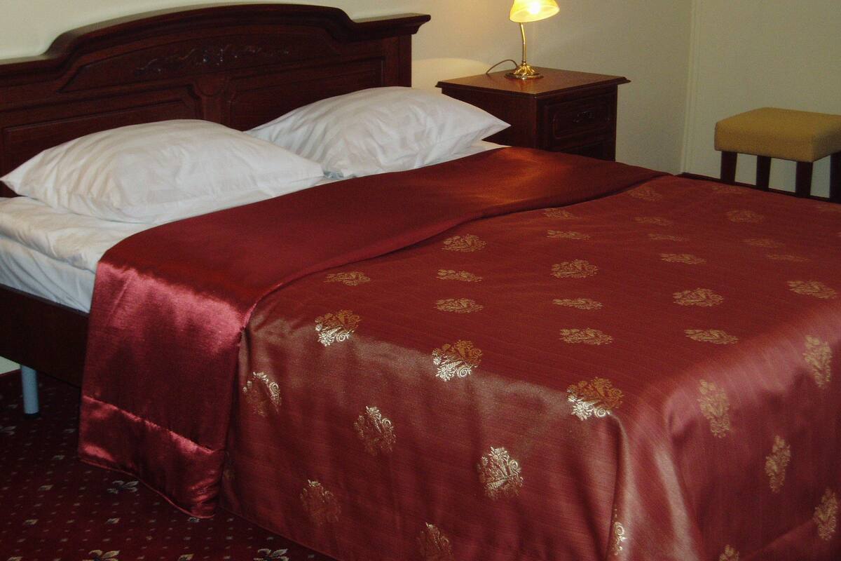 Una cama bien tendida con una colcha roja y almohadas blancas, una lámpara de noche y una mesita de madera.