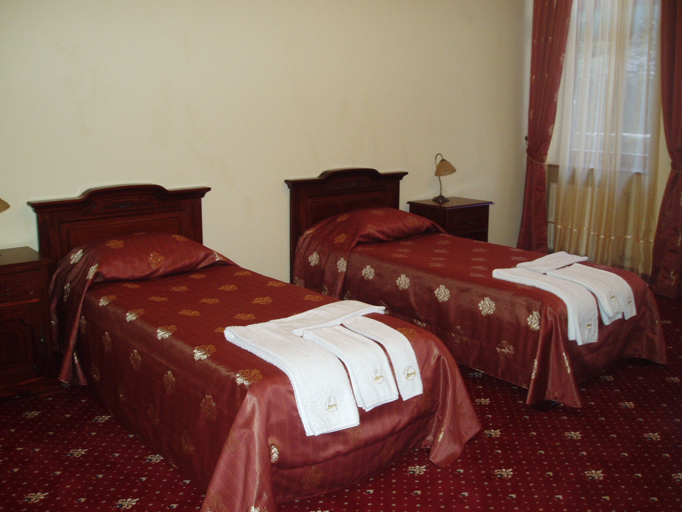 A hotel room with two single beds, each with a red bedspread and white towels. There is a wooden nightstand with a lamp between the beds, and a window with curtains to the right.