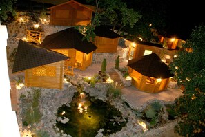 Une vue nocturne d’un chalet rustique doté de plusieurs cabanes en bois, d’un petit étang et de murs en pierre.