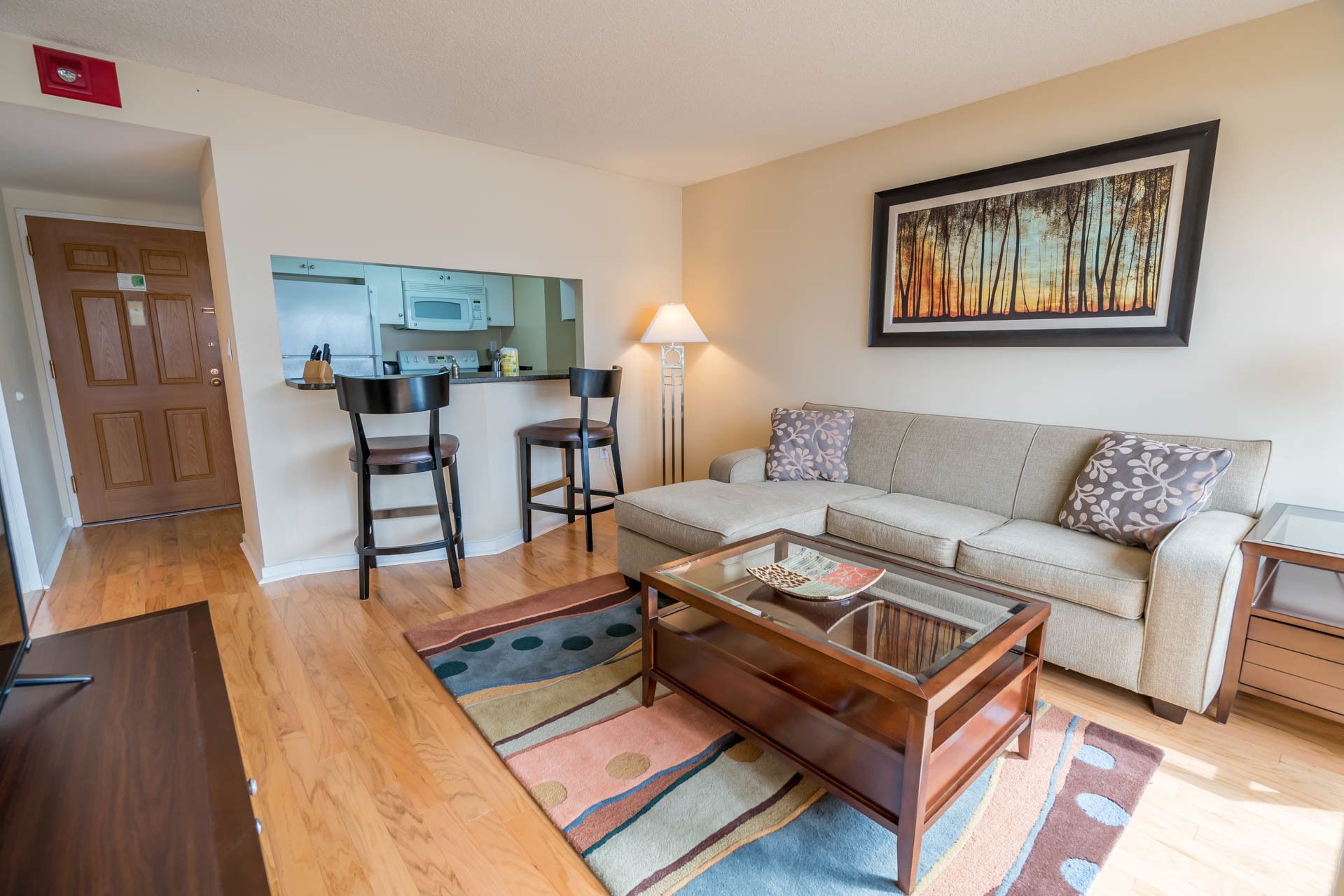 A living room with a sofa, two chairs, a coffee table, a kitchen area with a microwave, and a painting on the wall.