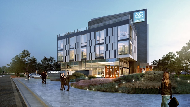 A modern hotel with a glass facade, a prominent AC logo, and a landscaped entrance area.