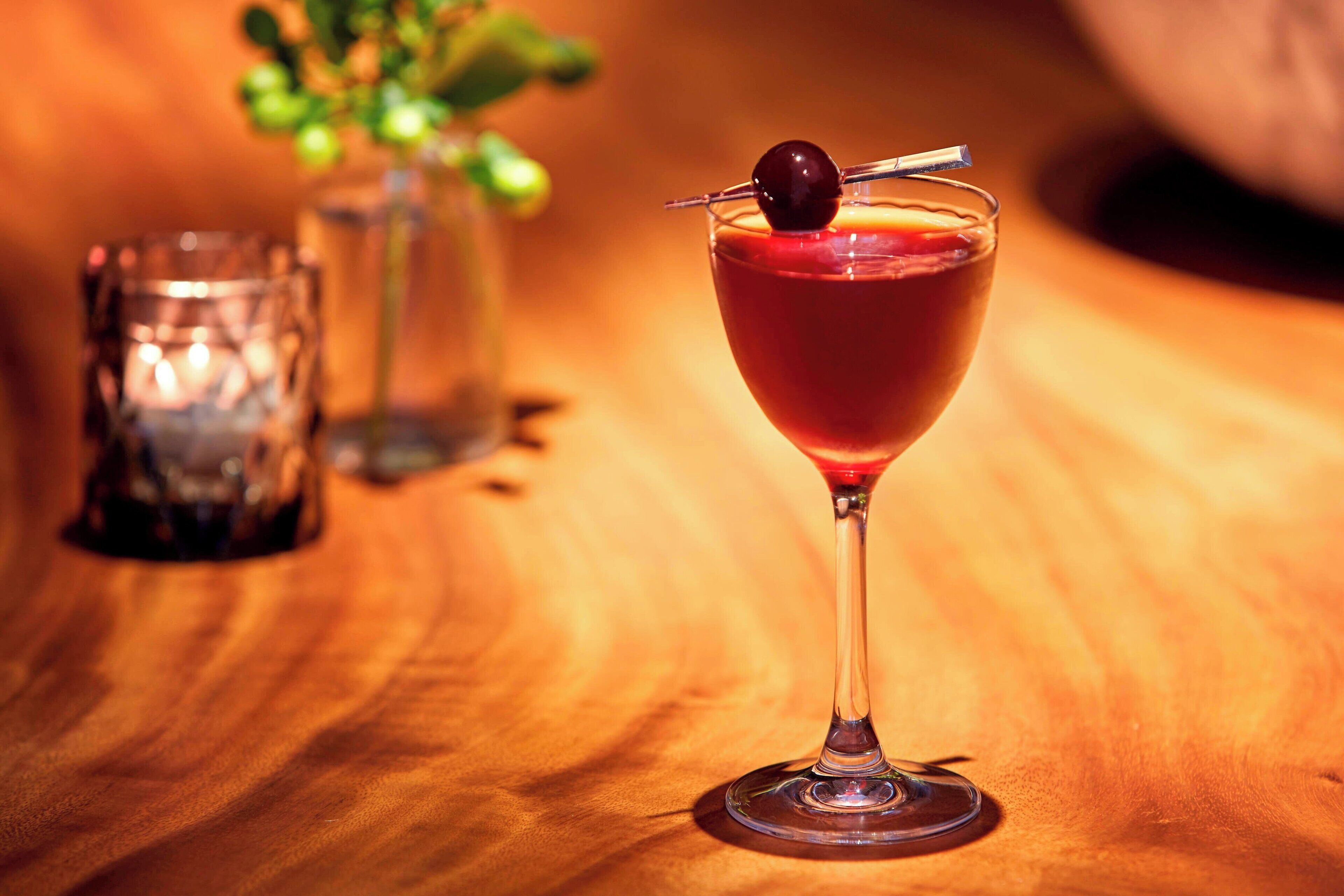 Un cocktail servi dans un verre à pied, décoré d’une cerise, sur une table en bois.