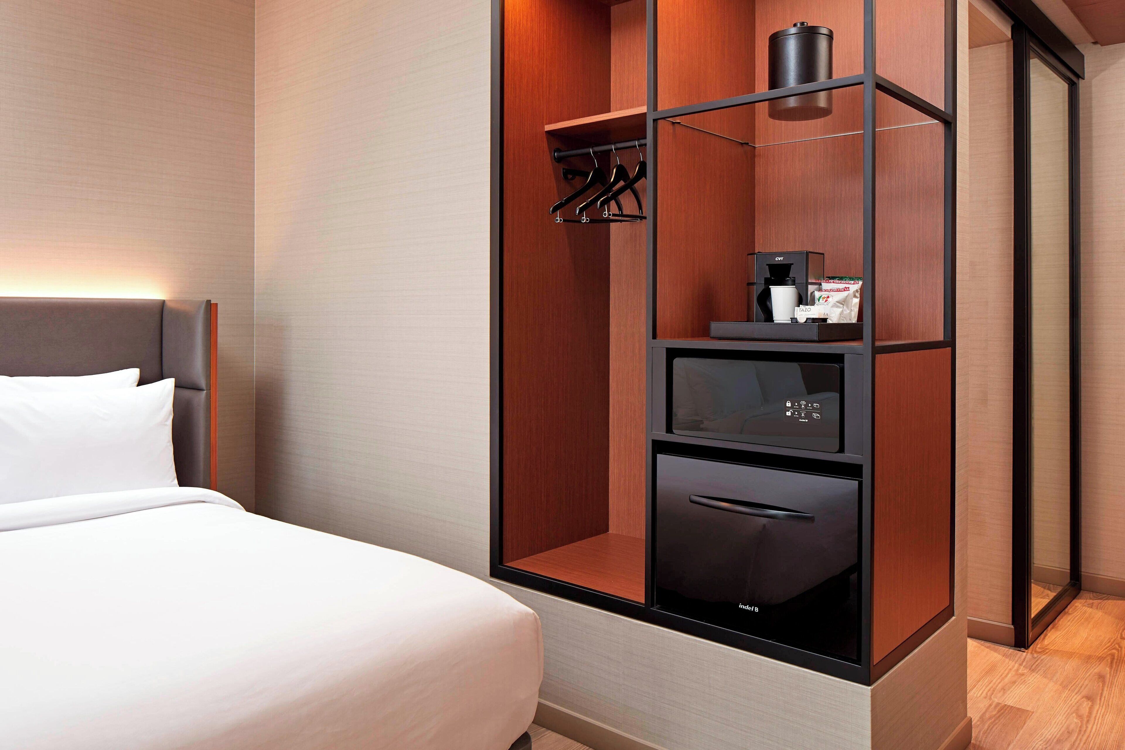 A modern hotel room with a bed, a built-in wardrobe with a coffee machine, and a small refrigerator.