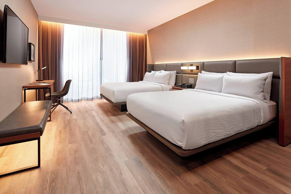 A hotel room with two beds, a desk, a chair, a TV, and a large window with curtains.