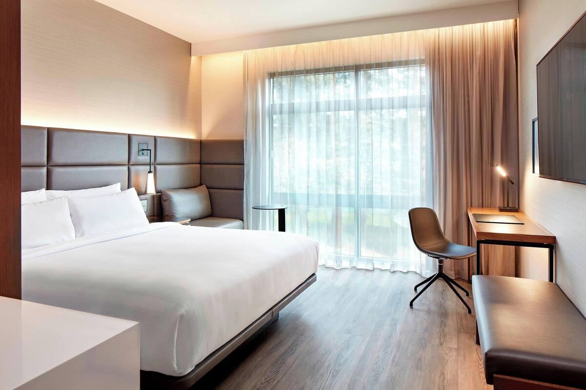 A modern hotel room with a large bed, a desk, a chair, and a bench.