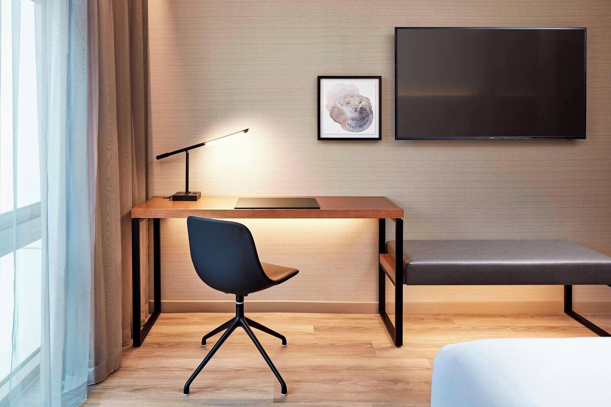 A modern hotel room with a wooden desk, a black office chair, a bench, a framed picture on the wall, and a flat-screen TV.