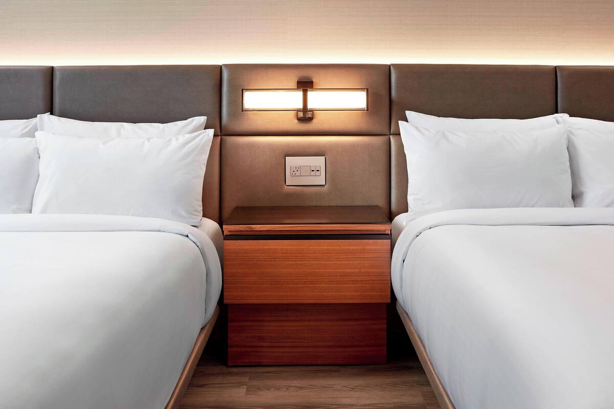 A hotel room with two beds, a nightstand with a lamp, and a power outlet.