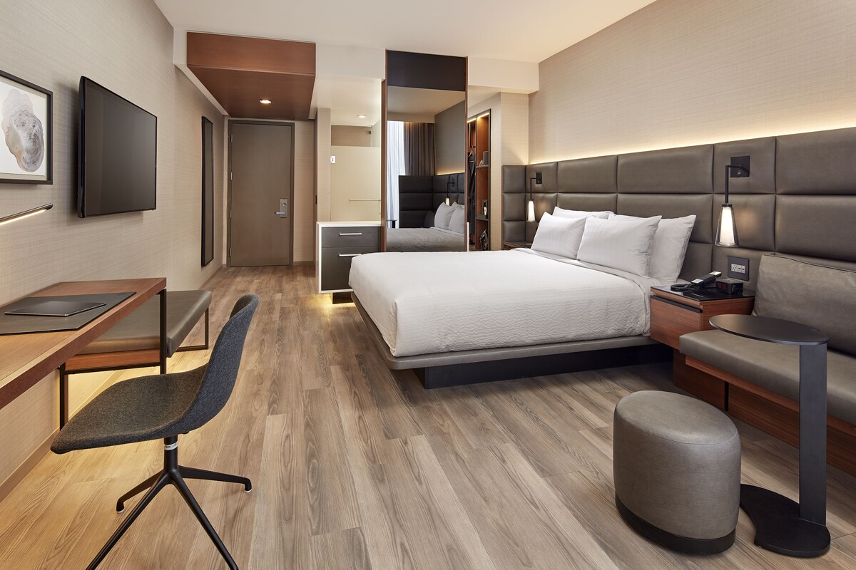 Une chambre d’hôtel moderne, dotée d’un grand lit, d’un bureau avec une chaise, d’une table de chevet et d’un petit pouf.