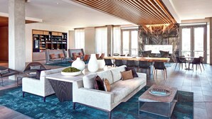 A spacious living area with a modern sofa, coffee table, and dining table. There is a bookshelf filled with books and decorative items. The room has large windows and a view of the city.