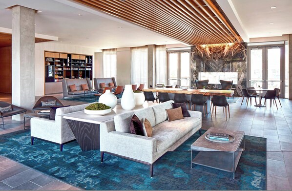 Amplia sala de estar con un sofá moderno, una mesa de centro y una mesa de comedor. Se aprecia una estantería llena de libros y objetos decorativos. La habitación cuenta con grandes ventanales y vistas a la ciudad.