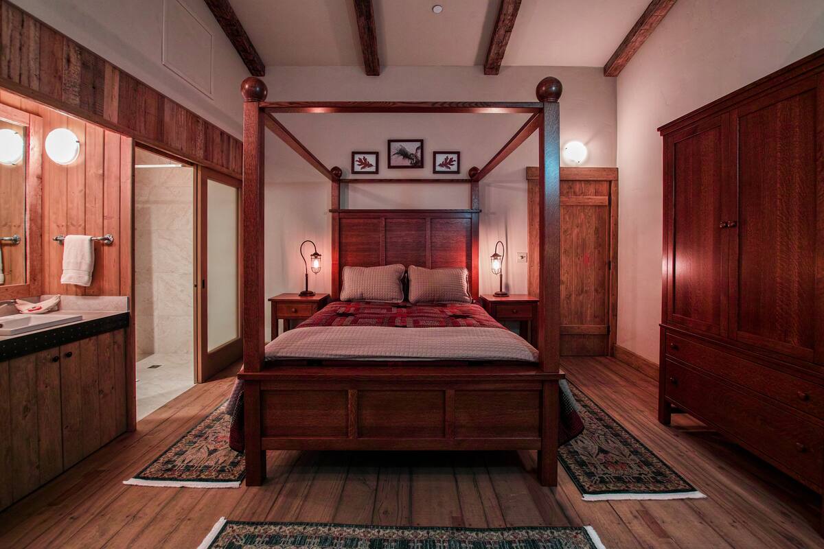 A four-poster bed with a wooden headboard, two bedside tables with lamps, a large wooden wardrobe, and a wooden floor.