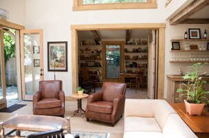 Amplia sala de estar con un sillón de cuero marrón, un sofá blanco y una mesa de centro de madera. Se observa una estantería llena de libros y objetos decorativos, además de un comedor con mesa y sillas de madera.