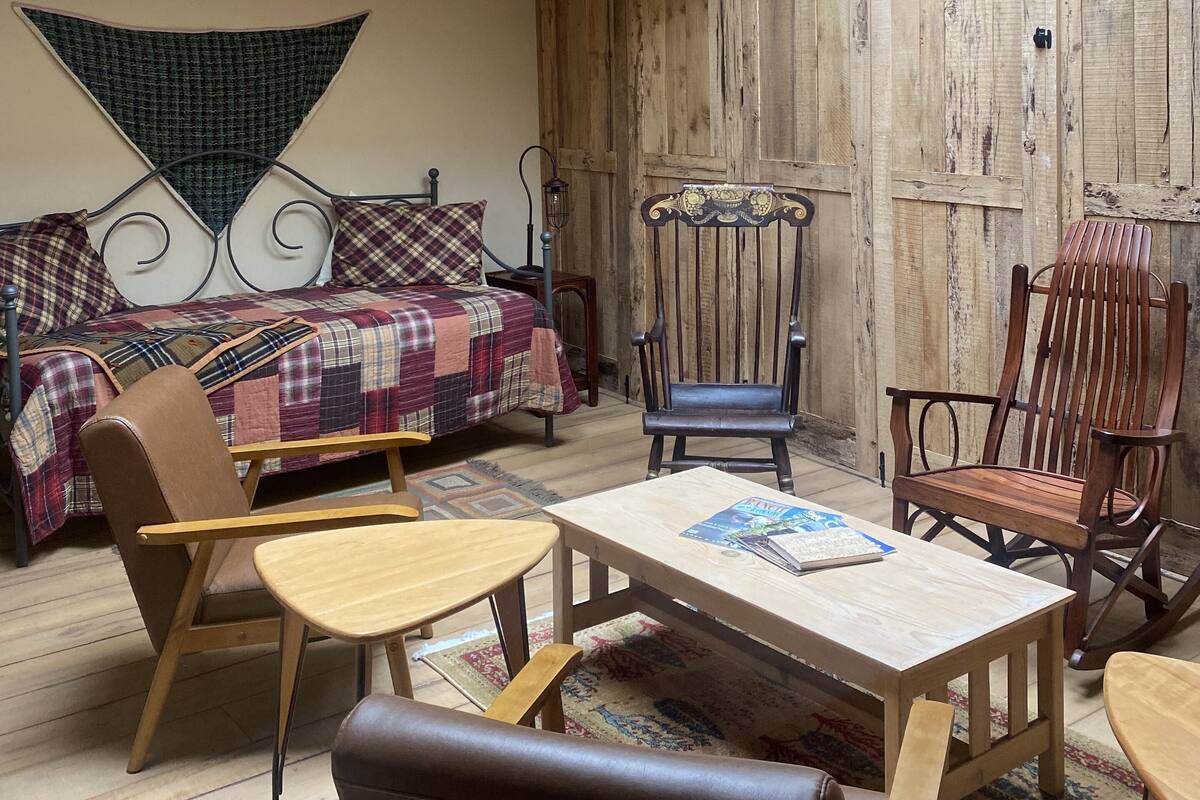 A cozy room with a sloped ceiling, wooden beams, a bed with a plaid blanket, two wooden chairs, a small table, and a patterned rug.